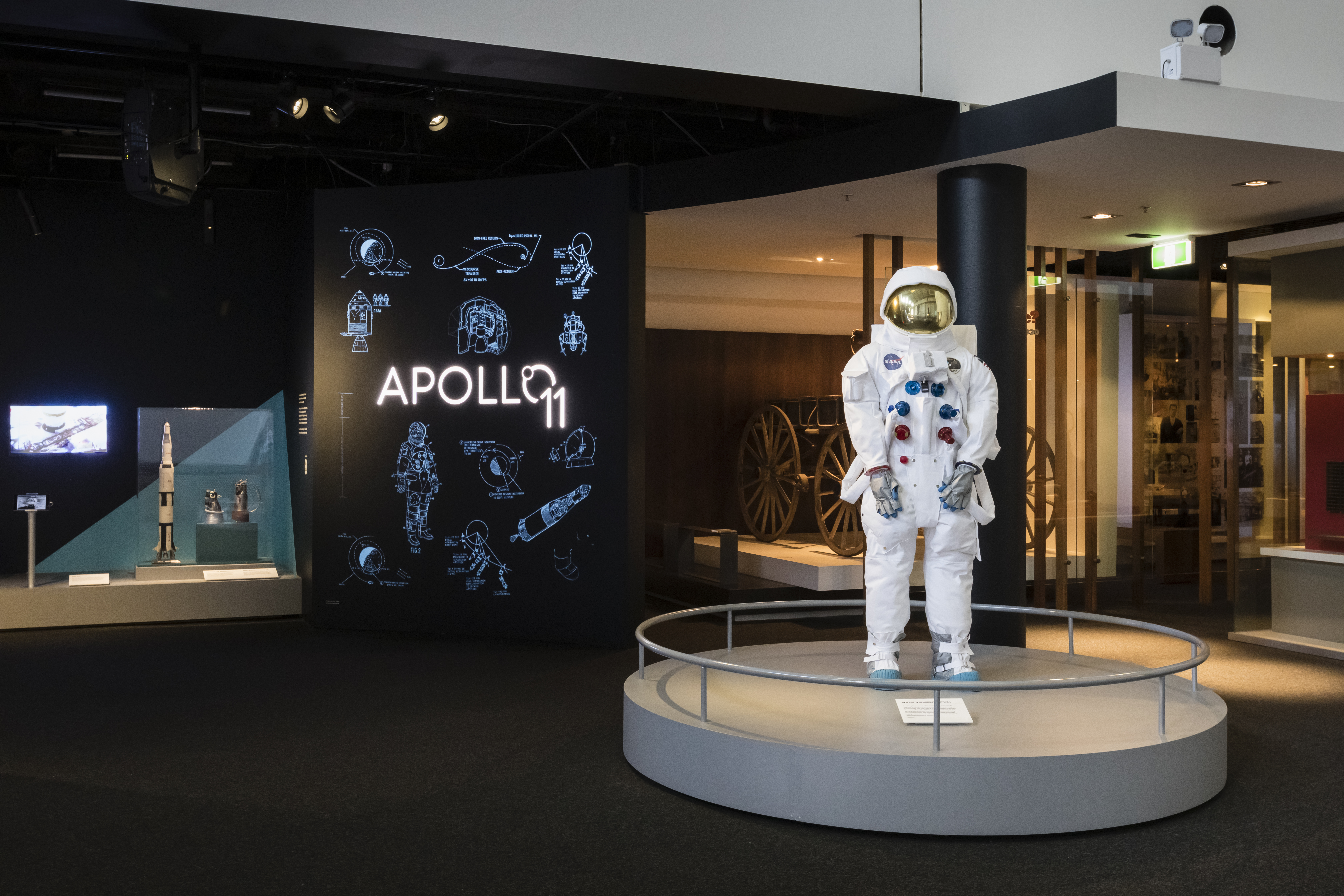 A mannequin wearing a space suit stands on a silver circular plinth. To the left is a title wall reading 'Apollo 11' with diagrams of spacecraft around it.